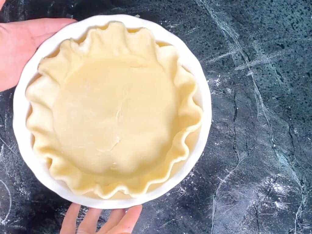 A 9-inch pie dish lined with rolled Crisco pie crust dough on a warm wood counter, with a rolling pin and a small bowl of flour nearby