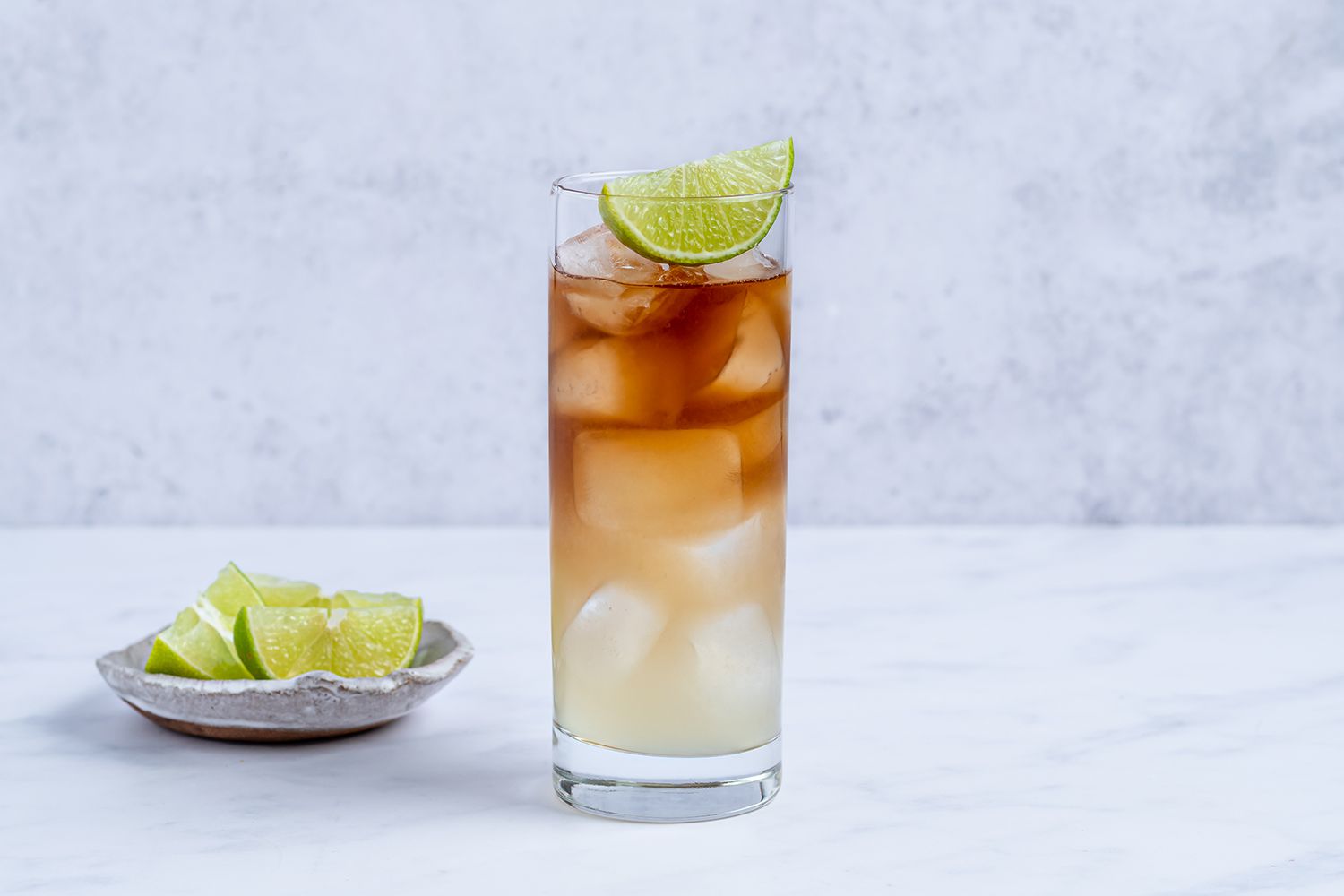 A Dark and Stormy cocktail in a tall glass with lots of ice, a dark rum float layered over ginger beer, and a lime wedge on the rim on a softly lit kitchen counter