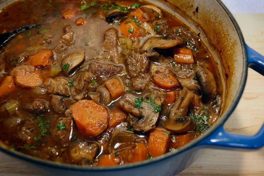 A Dutch oven filled with beef bourguignon, showing tender beef chunks in a glossy red wine sauce with mushrooms and pearl onions, on a warm kitchen counter with soft natural light