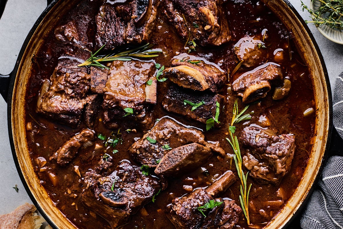 A Dutch oven filled with fall-apart beef short ribs in a glossy red wine gravy, garnished with herbs on a warm kitchen counter