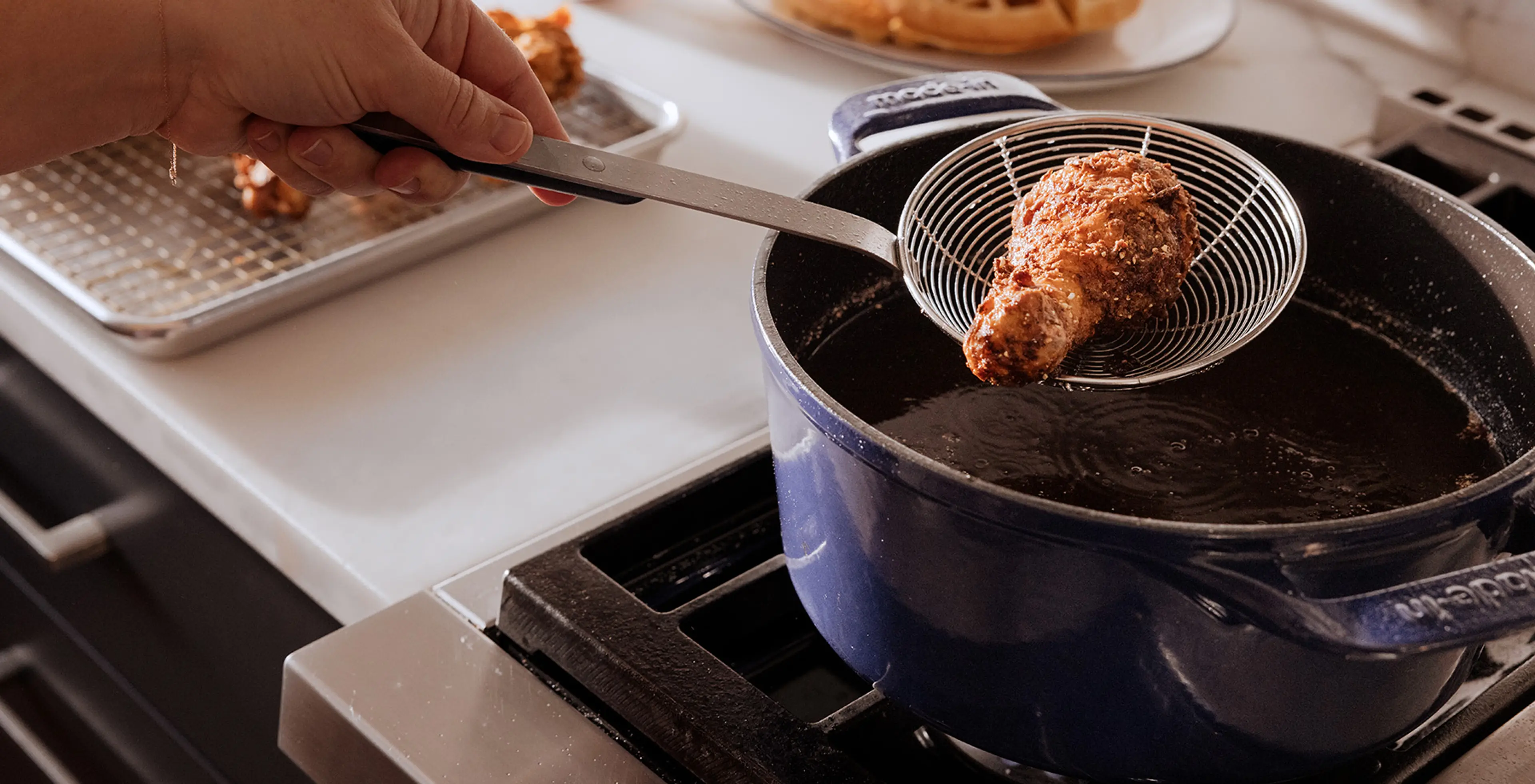 A Dutch oven on a stovetop filled with hot oil while a spider strainer lifts pale first-fry potatoes above the pot