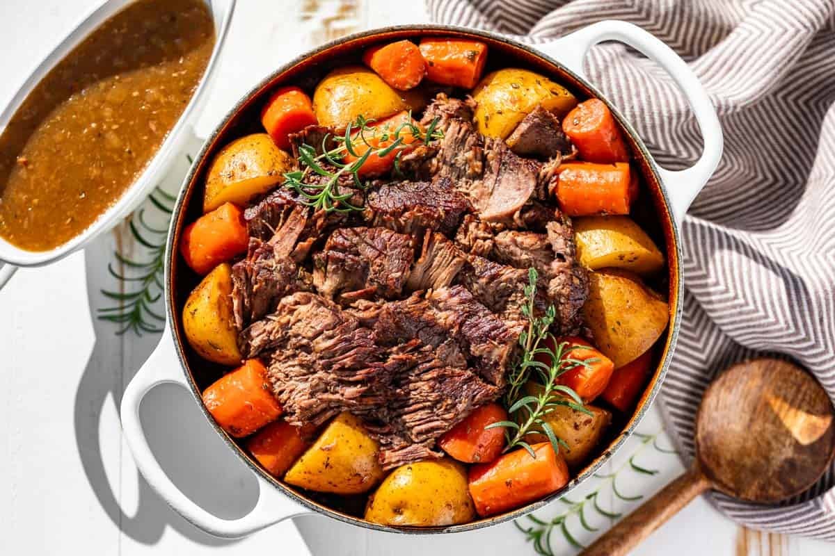 A Dutch oven pot roast sliced and piled on a platter with carrots and potatoes, coated in glossy brown gravy on a rustic wooden table
