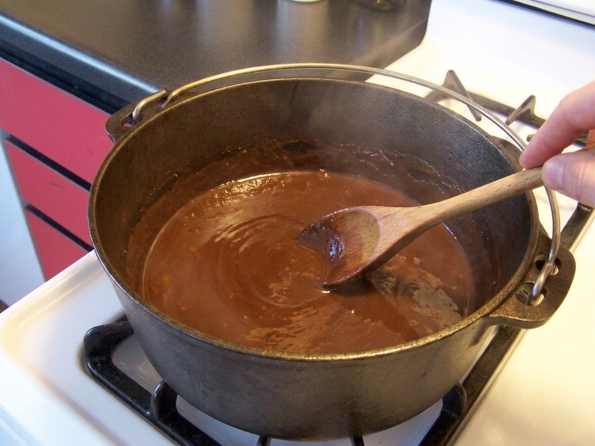 A Dutch oven with a smooth, glossy dark brown roux being stirred with a wooden spoon on a stovetop