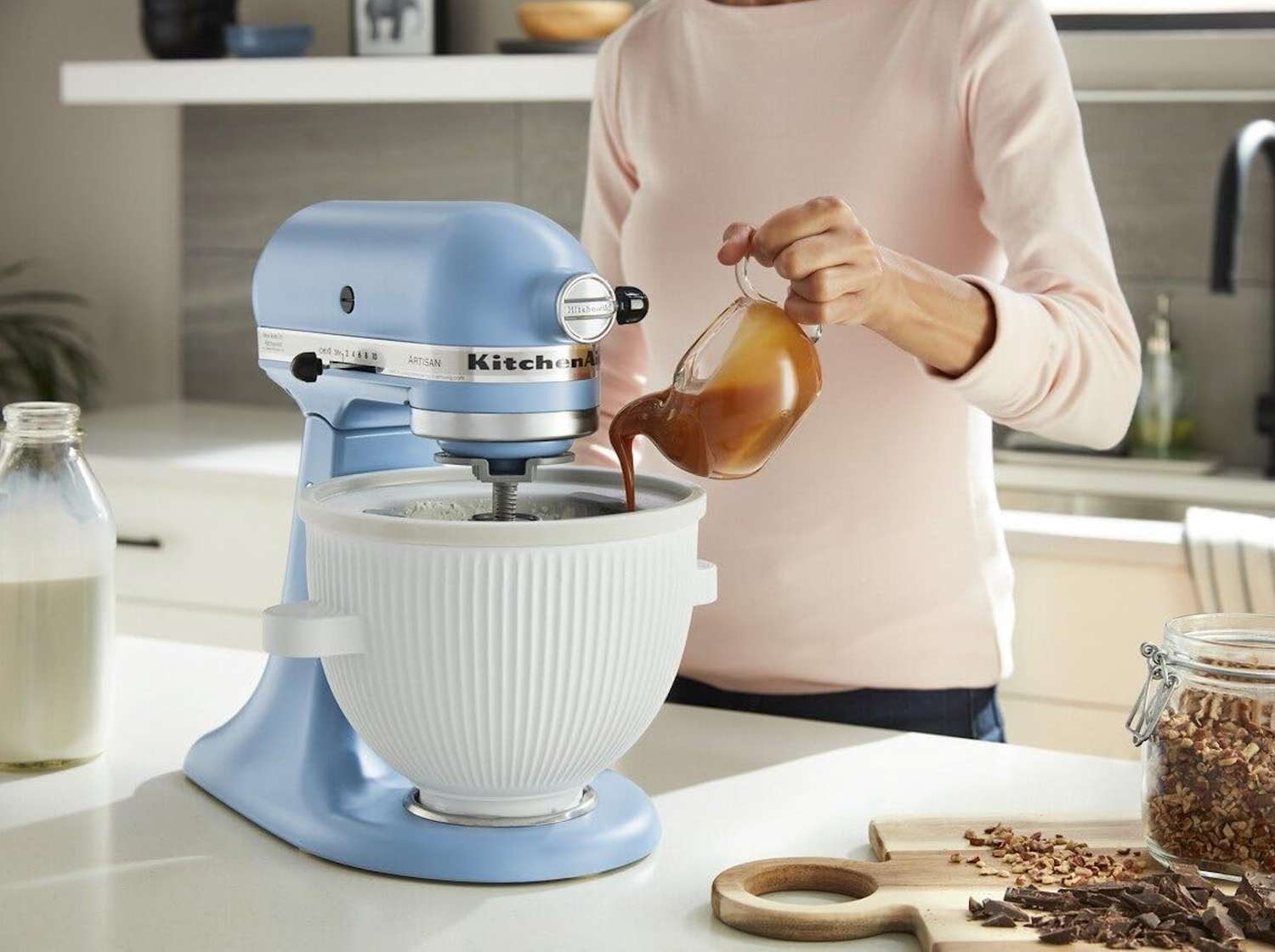 A KitchenAid stand mixer fitted with the ice cream maker attachment on a bright kitchen counter, with a bowl of freshly churned vanilla ice cream nearby