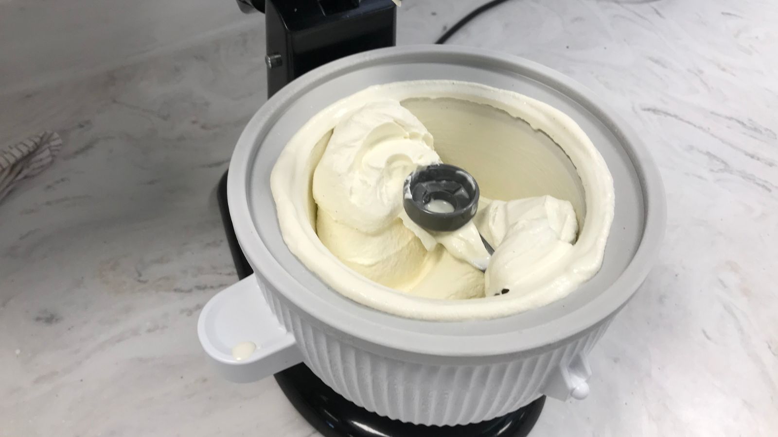 A KitchenAid stand mixer running with the ice cream maker attachment while pale vanilla ice cream thickens inside the frozen bowl