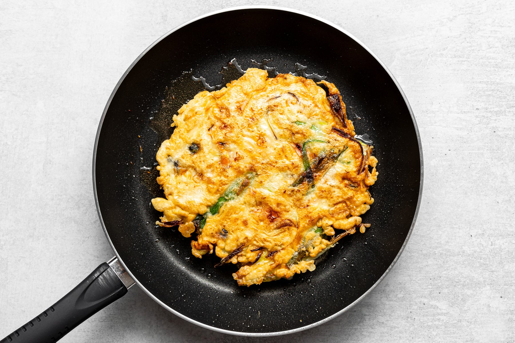 A Korean scallion pancake being flipped in a skillet with a wide spatula over a home stove