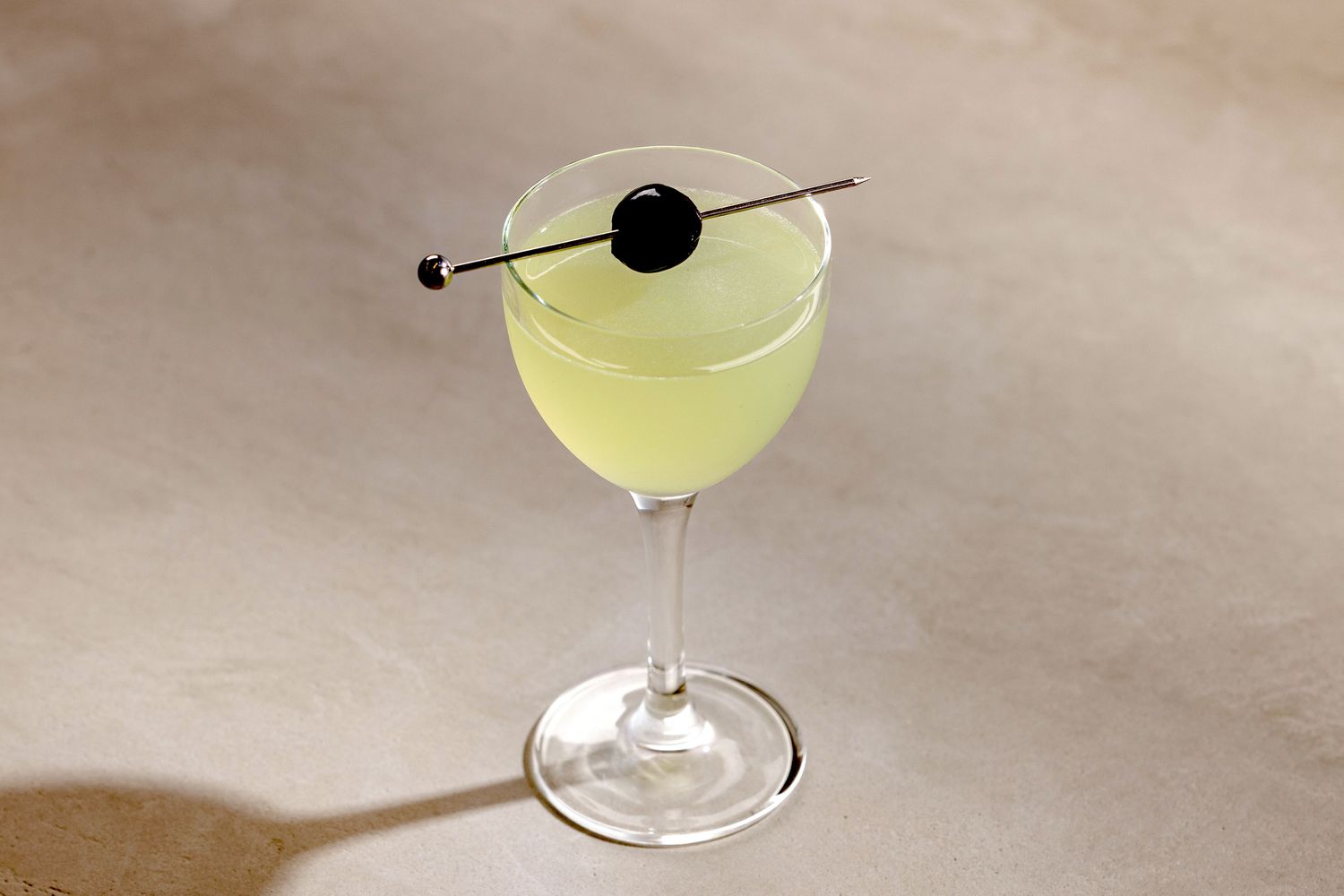 A Last Word cocktail in a chilled coupe glass on a light stone kitchen counter, pale green drink with a lime twist, soft natural window light