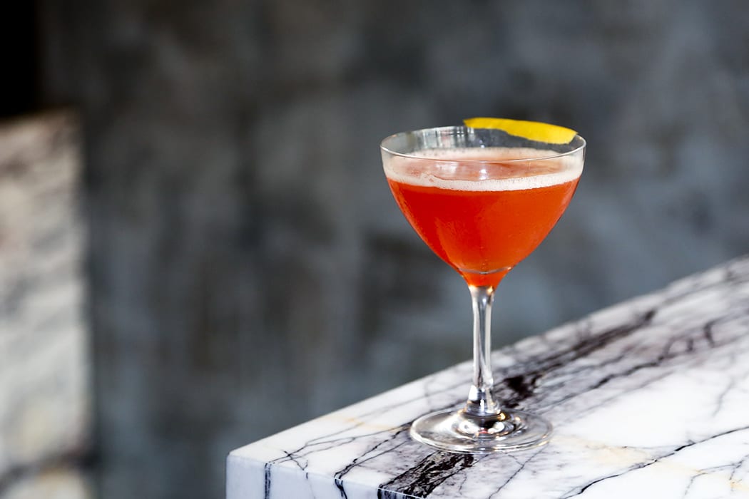 A Paper Plane cocktail in a chilled coupe glass with a thin lemon twist on a light stone countertop under warm natural kitchen light