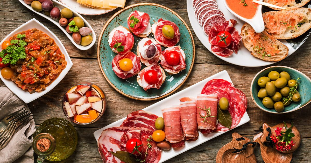 A Spanish tapas spread on a wooden table with patatas bravas, garlic shrimp, pan con tomate, marinated olives, and toasted almonds