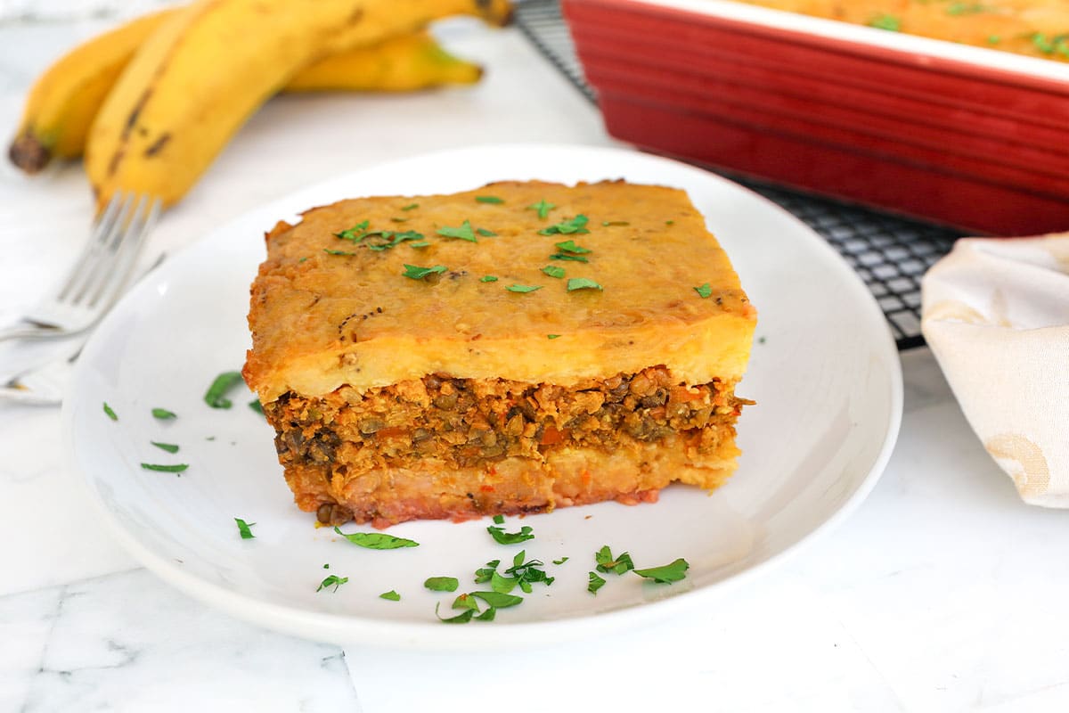 A baked Puerto Rican pastelón in a ceramic baking dish with golden sweet plantain slices on top and a corner slice lifted out to show layers of beef picadillo and melted cheese