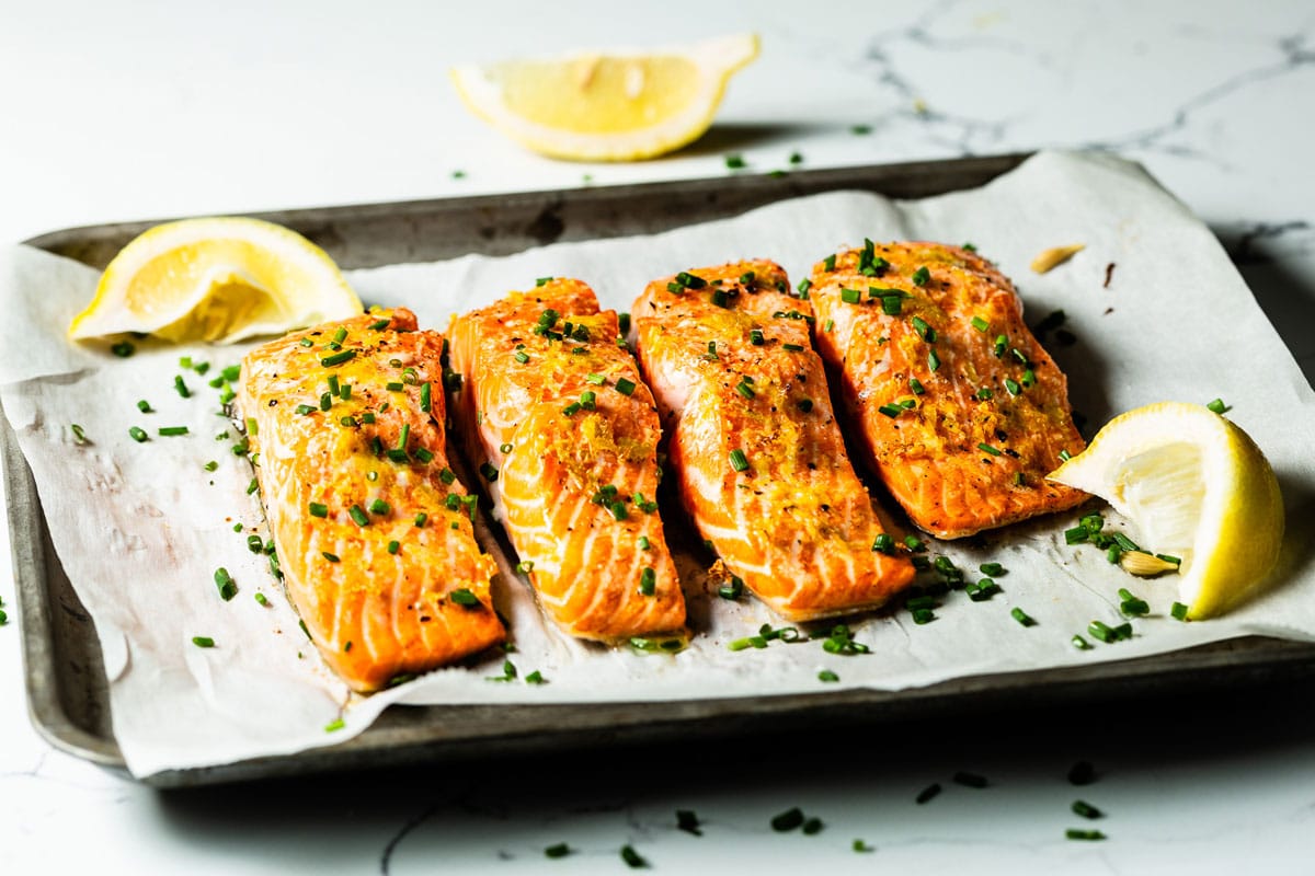 A baked salmon fillet on a sheet pan with lemon slices and herbs, lightly browned edges, photorealistic food photography