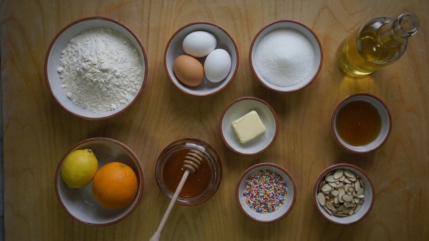 A baking setup on a kitchen counter with flour, eggs, sugar, butter, lemons, orange, honey, sprinkles, and sliced almonds arranged in small bowls