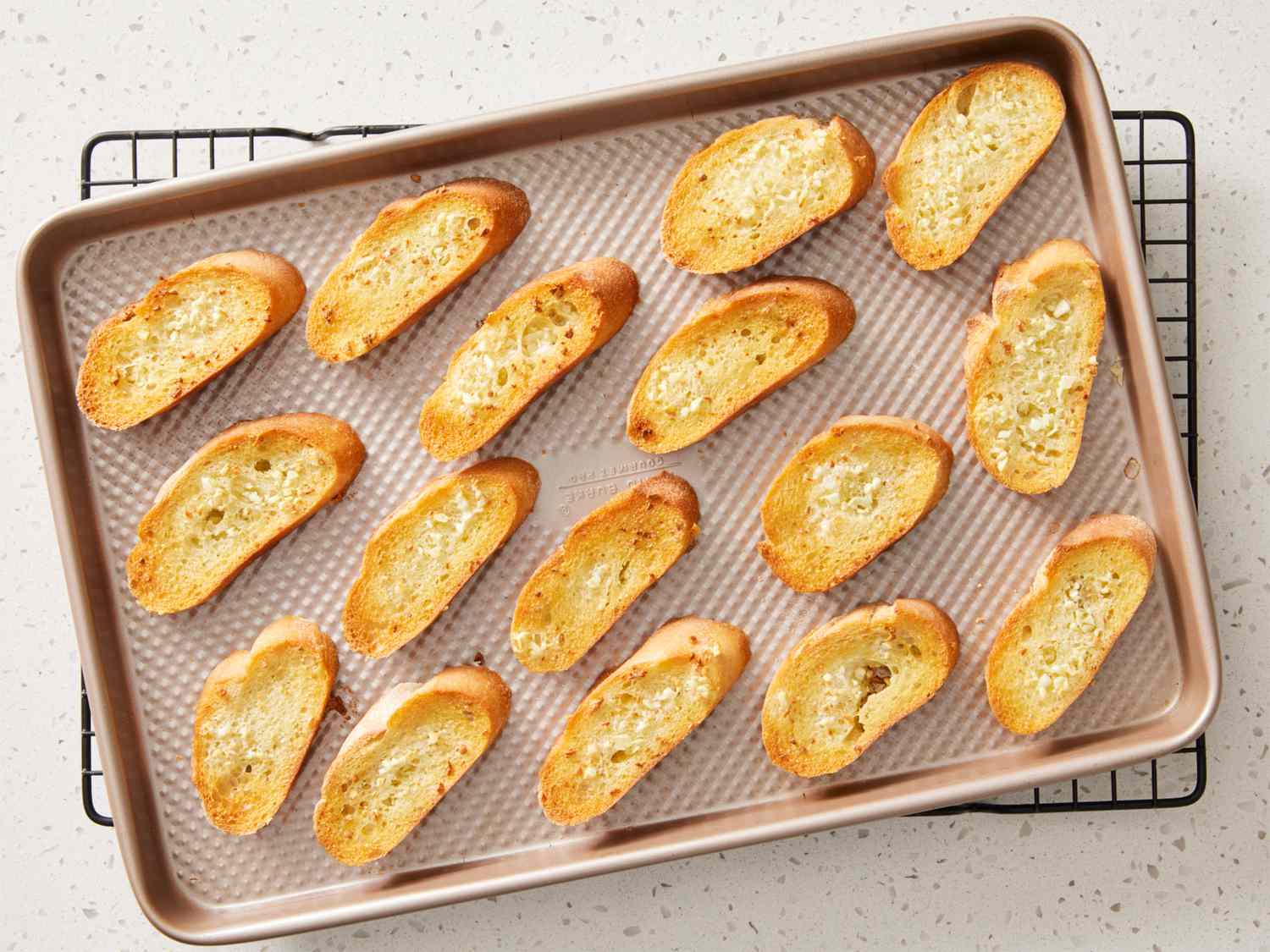 A baking sheet filled with evenly toasted golden crostini rounds cooling on a kitchen counter under warm light