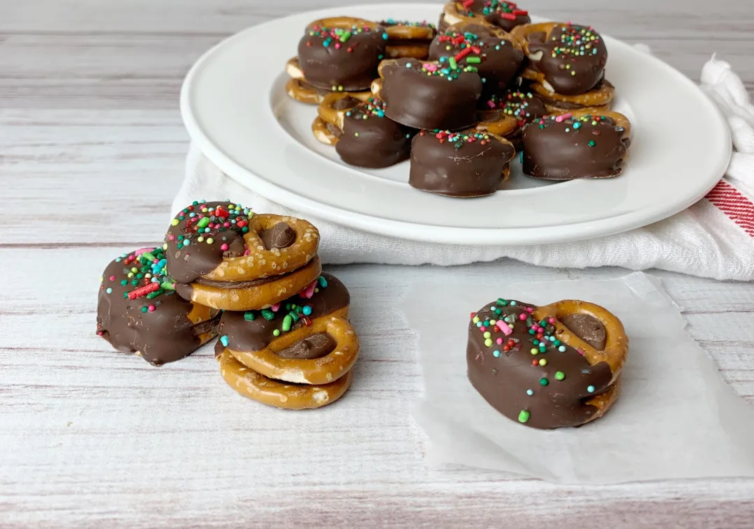 A baking sheet filled with pretzel Rolo bites, each mini pretzel topped with a softened chocolate caramel candy and a pecan half, on a parchment-lined pan in a warm home kitchen