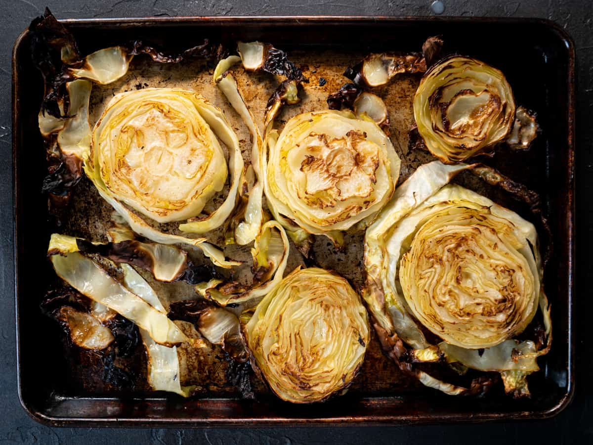 A baking sheet holding roasted cabbage steaks with deeply browned edges, sprinkled with herbs and lemon zest, set on a warm-toned kitchen counter
