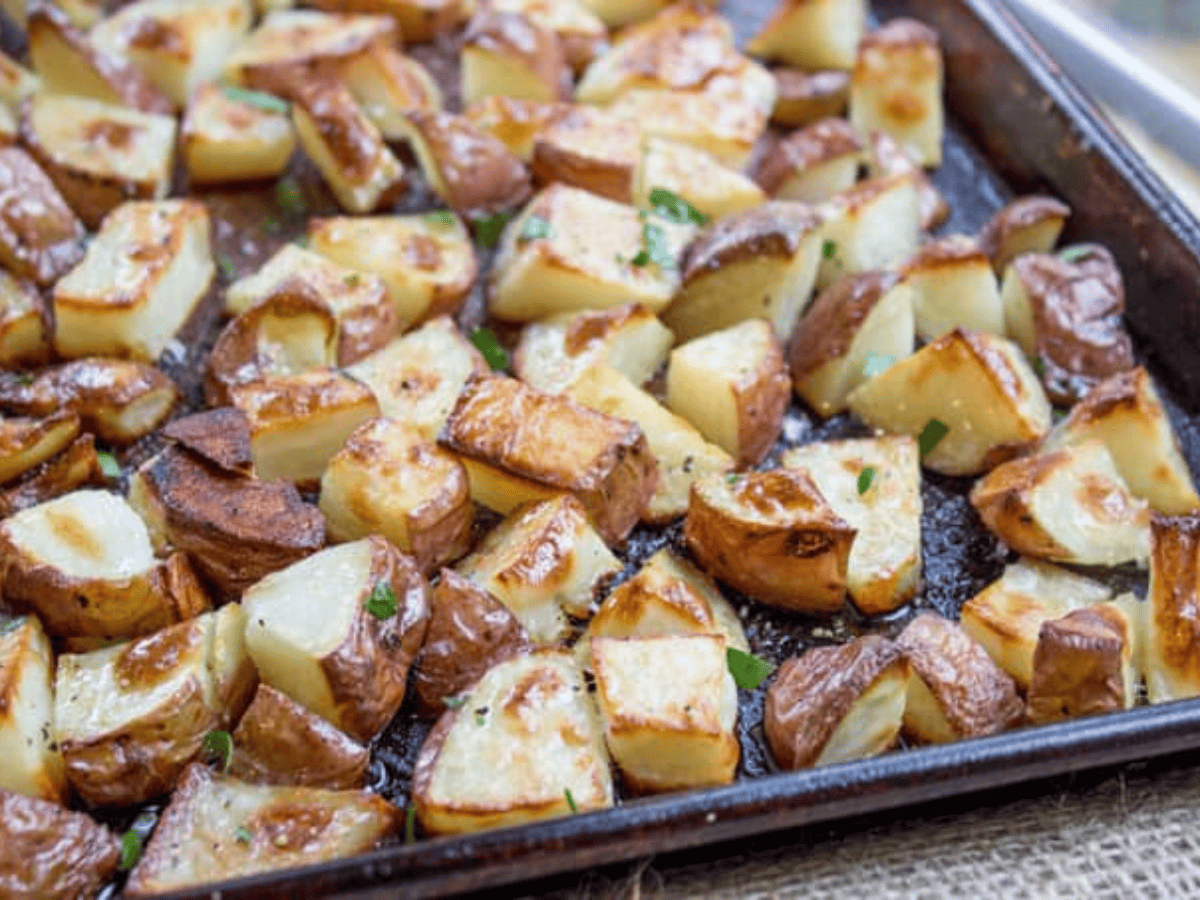A baking sheet of roasted scarlet potatoes with crisp browned edges, scattered rosemary, and a glossy lemon butter sheen on a warm kitchen counter