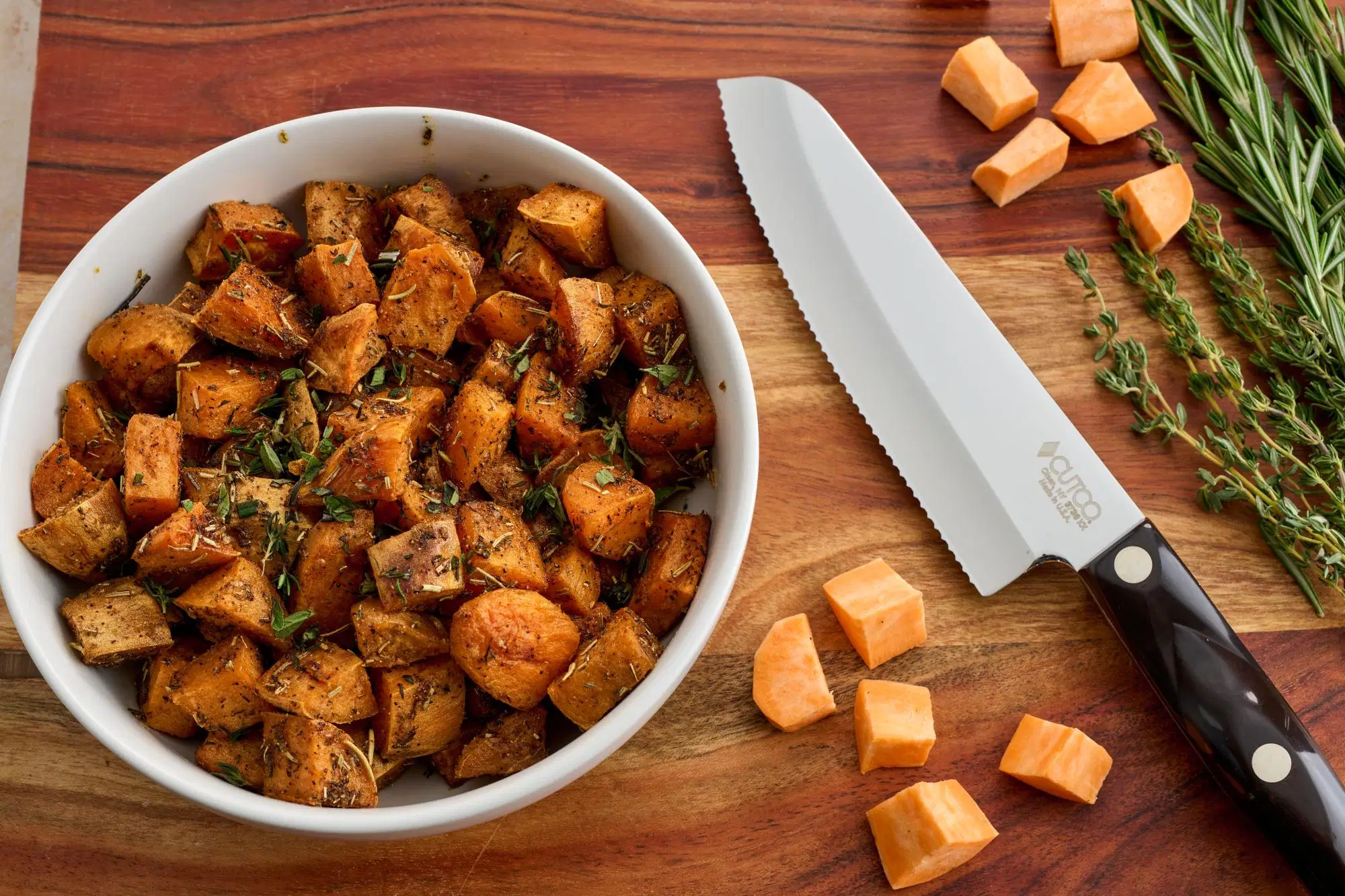 A baking sheet of roasted sweet potato cubes with deeply browned edges and scattered herbs, shot on a warm kitchen counter with soft natural light
