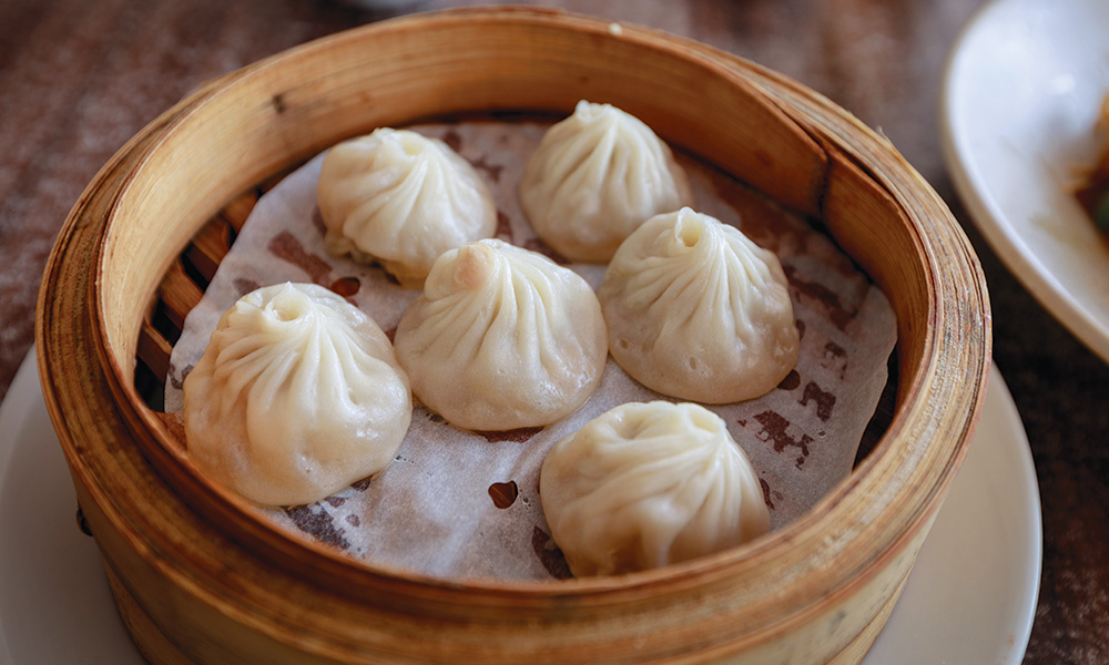 A bamboo steamer basket filled with freshly steamed siu long bao soup dumplings lined on parchment, with a small dish of black vinegar and ginger on the side on a warm kitchen table