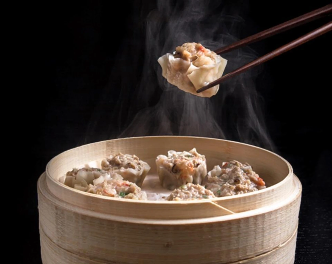 A bamboo steamer basket filled with pork and shrimp shumai, each topped with a small dot of orange roe, set on a wooden table with dipping sauce nearby
