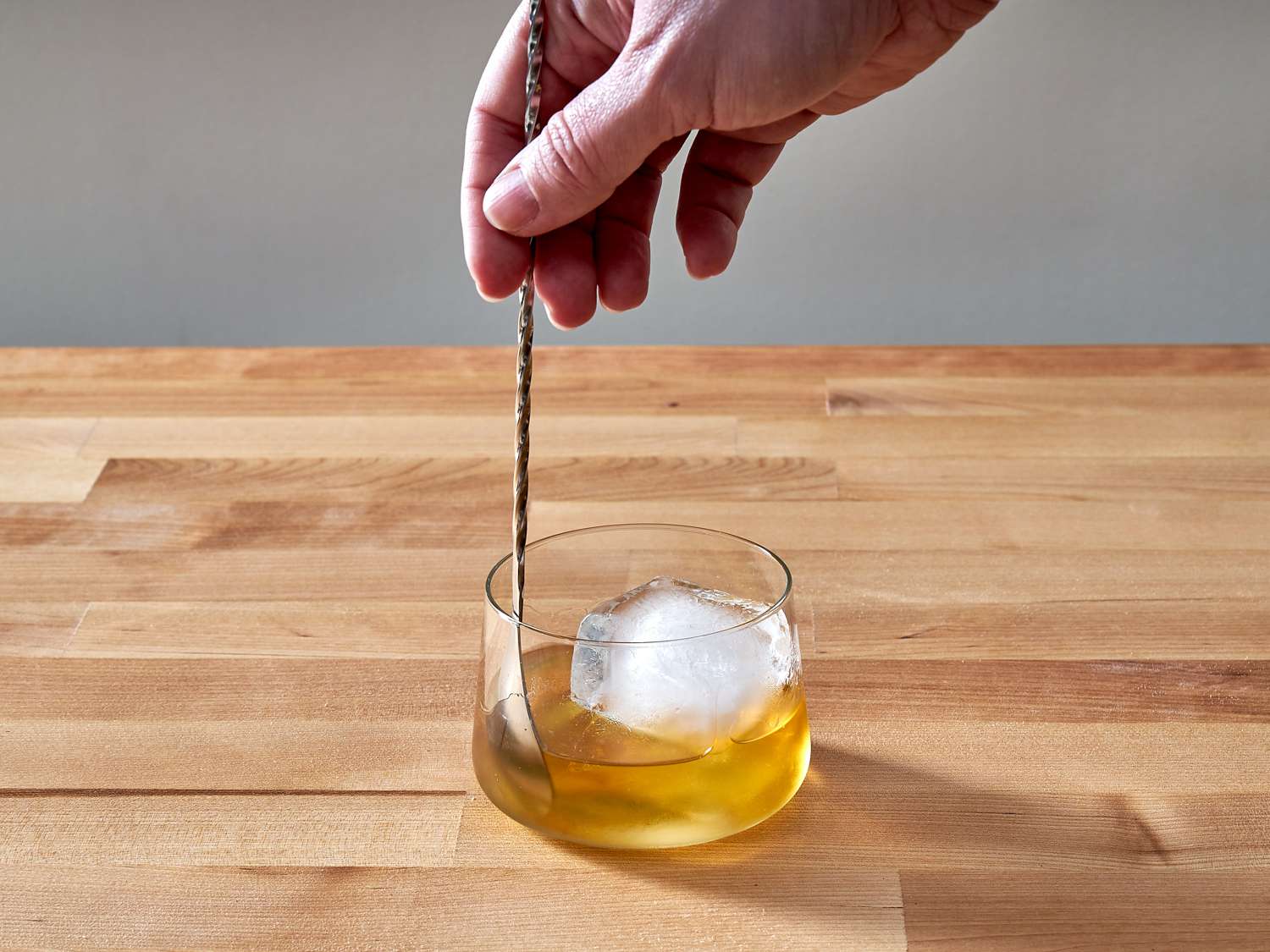 A bar spoon stirring a Rusty Nail cocktail in a rocks glass over a large ice cube on a kitchen counter with warm lighting