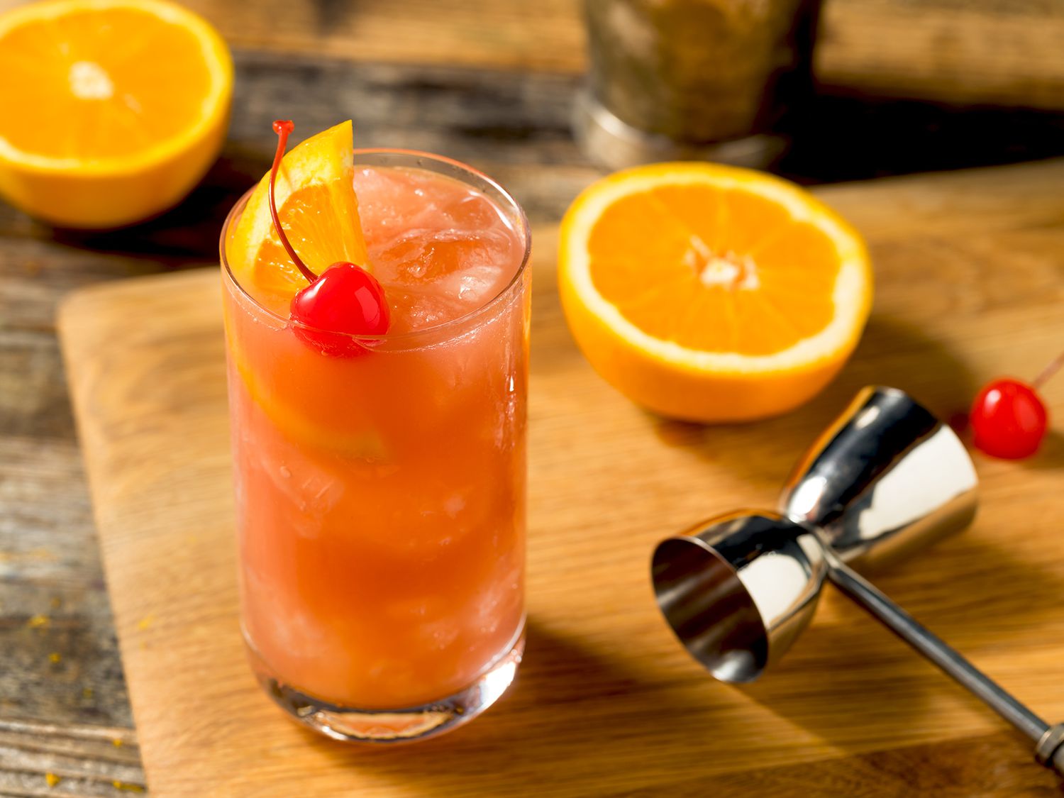A bar spoon stirring a pink and orange cocktail in a tall glass filled with ice on a clean kitchen counter