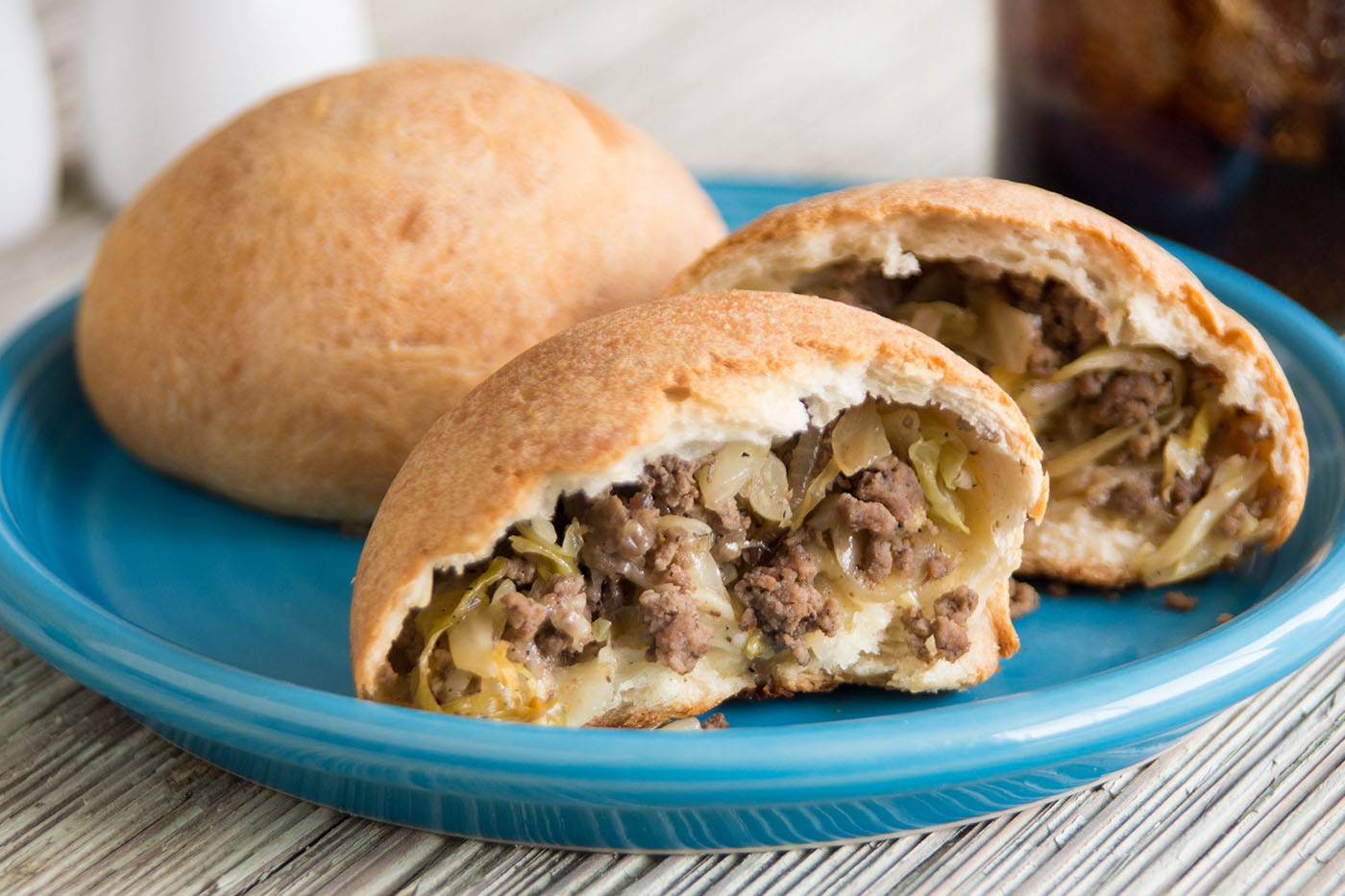 A basket lined with a towel holding several golden-brown bierocks, one split open to show the beef and cabbage filling, warm kitchen table setting