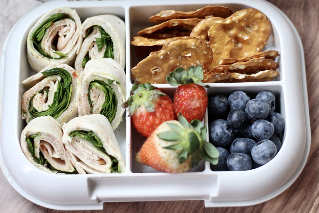 A bento lunchbox with turkey pinwheels, cheddar cubes, cucumber slices, strawberries, pretzels, and a small cup of ranch dip