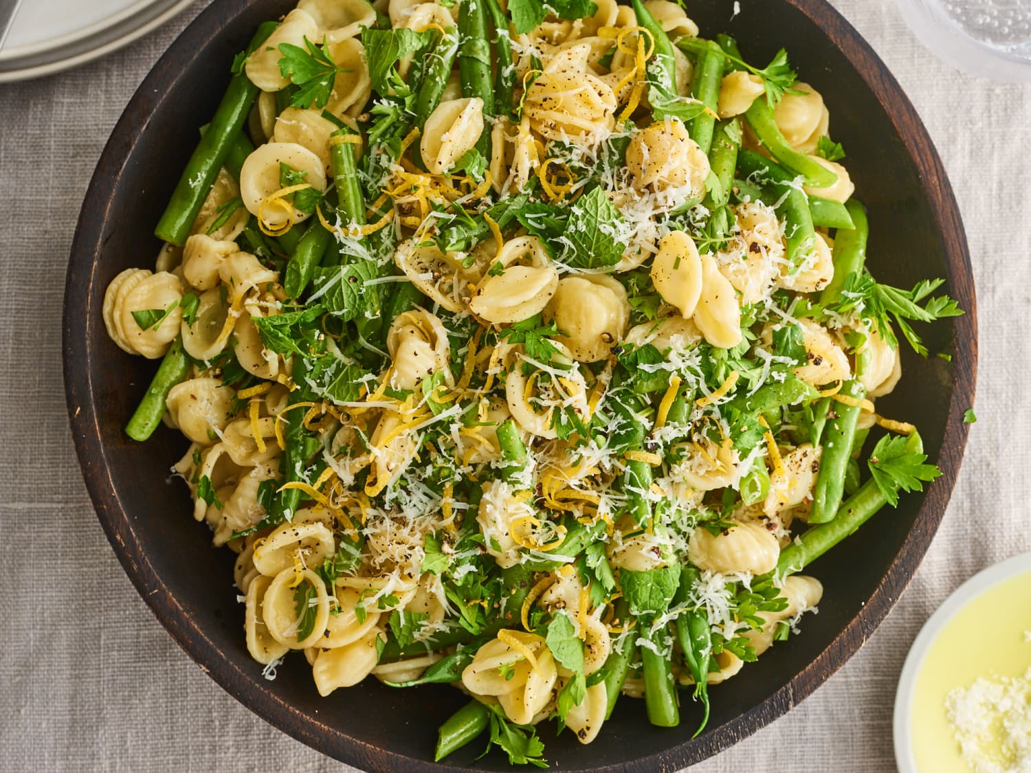 A big serving bowl of lemony pasta salad with cherry tomatoes, cucumbers, chickpeas, fresh herbs, and crumbled feta on a light kitchen counter
