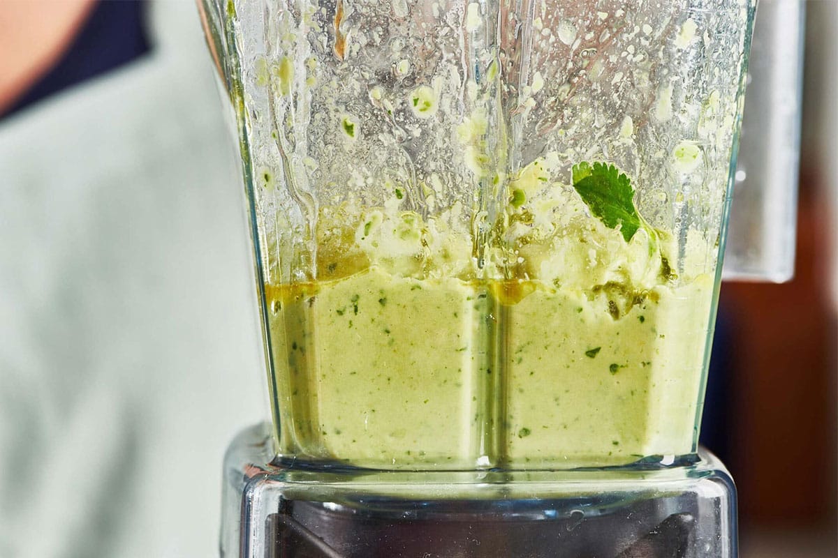 A blender cup filled with avocado, herbs, yogurt, lime juice, and olive oil before blending on a kitchen counter