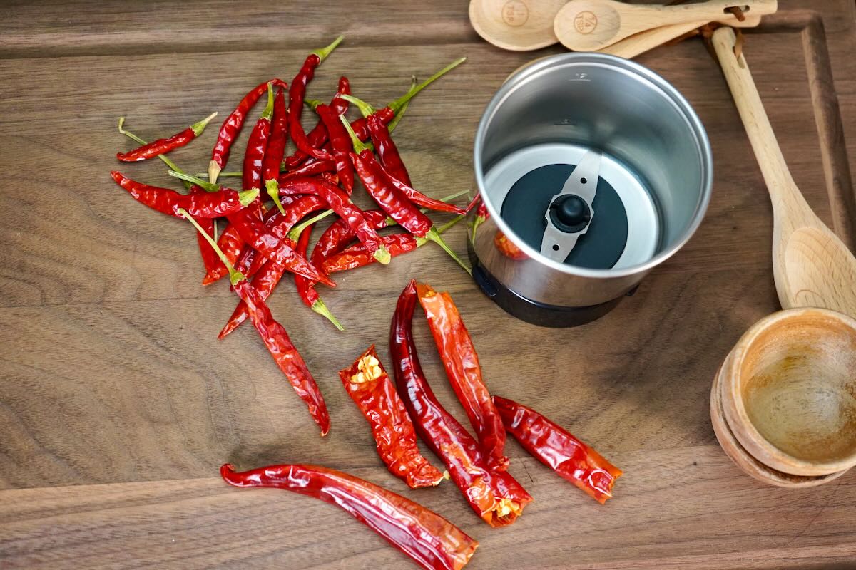 A blender cup filled with toasted dried chiles and spices ready to be blended into chili powder on a kitchen counter