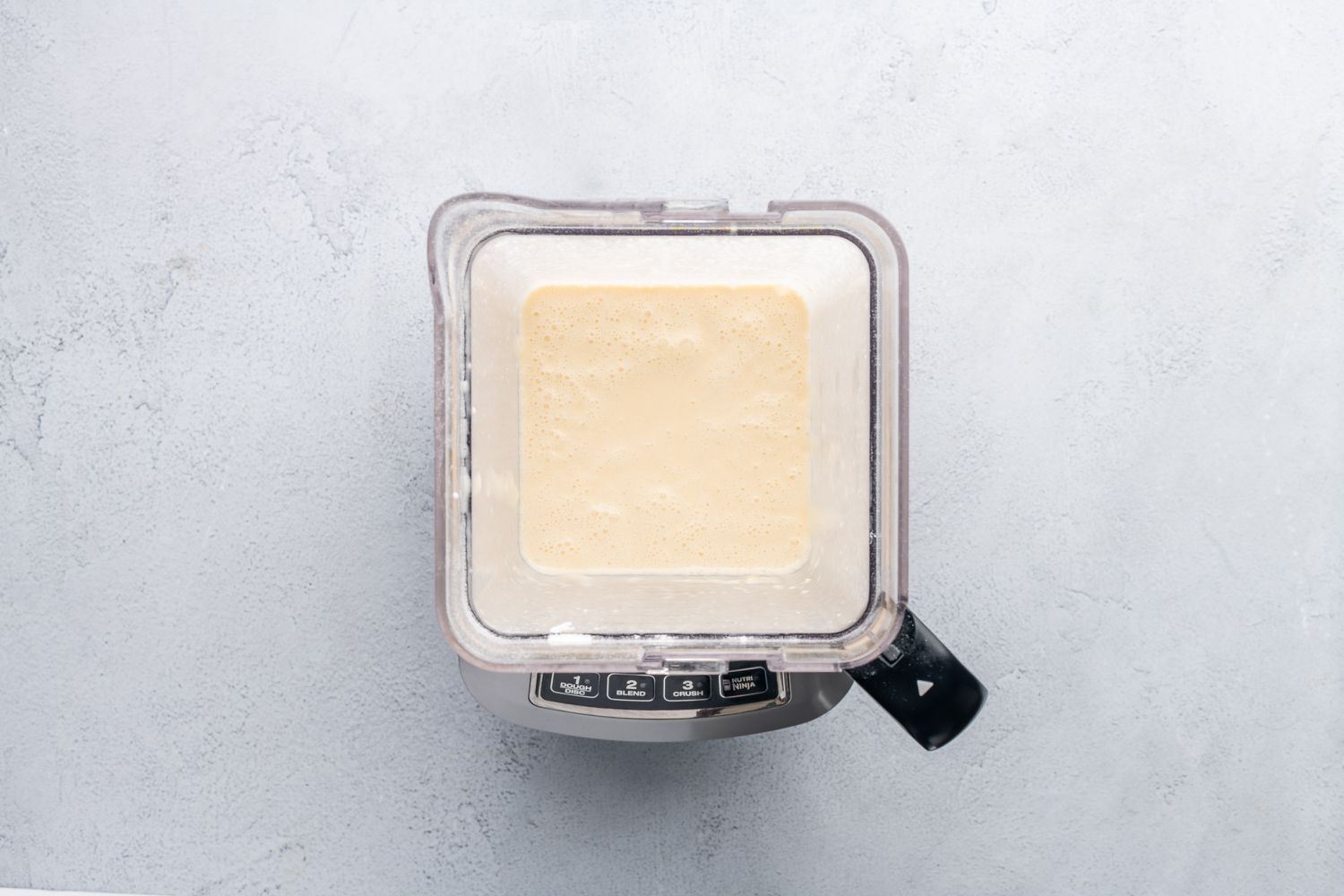 A blender jar filled with pale crepe batter on a kitchen counter with the lid ready to go on