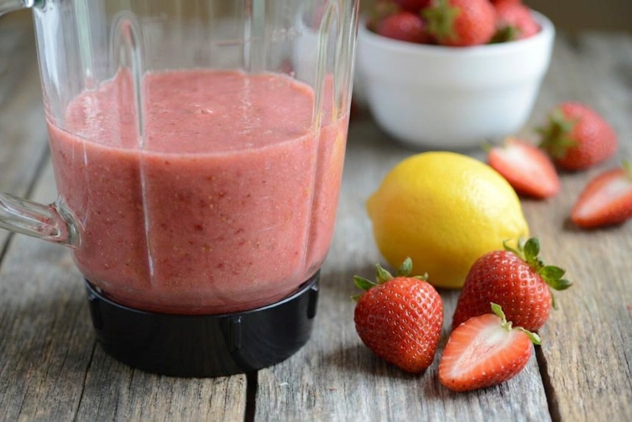 A blender jar filled with pink berry and applesauce puree on a kitchen counter next to fresh berries and a lemon