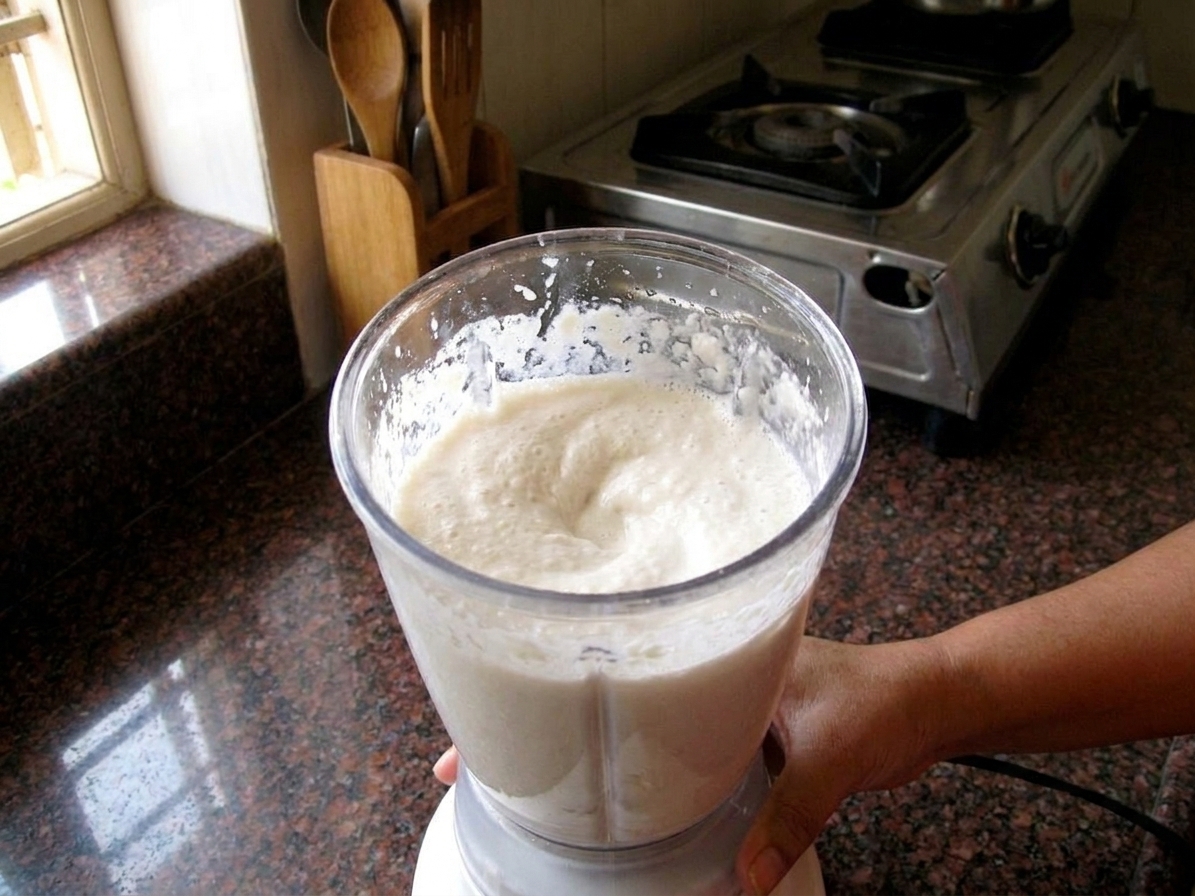 A blender jar filled with thick, pale urad dal batter mid-blend on a kitchen counter