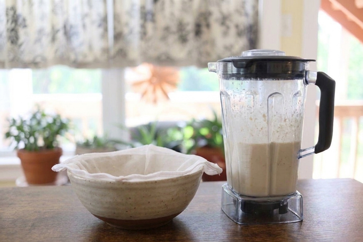 A blender next to a nut milk bag set over a large bowl on a bright kitchen counter, ready for straining oat milk