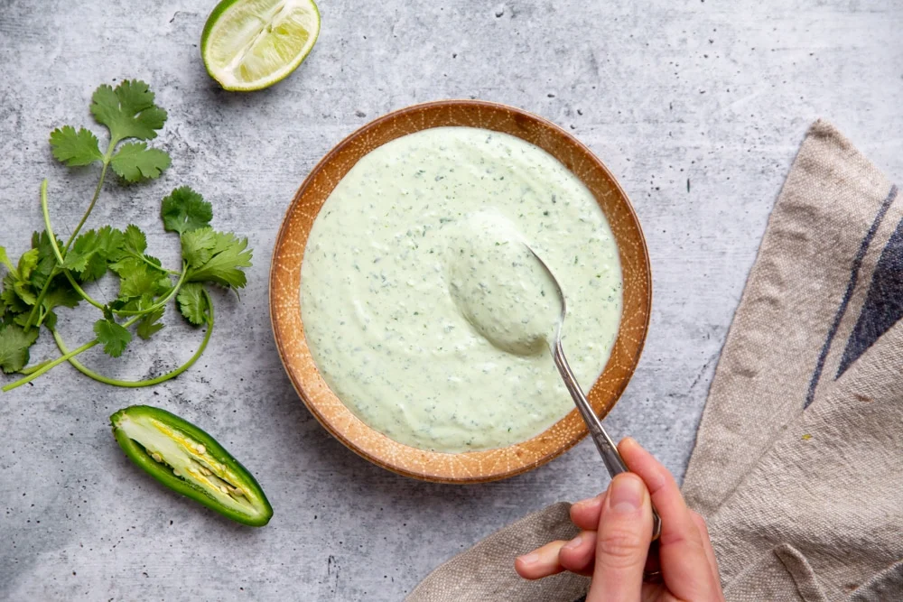A blender on a kitchen counter blending a creamy green basil lime sauce with a hand holding the lid in place