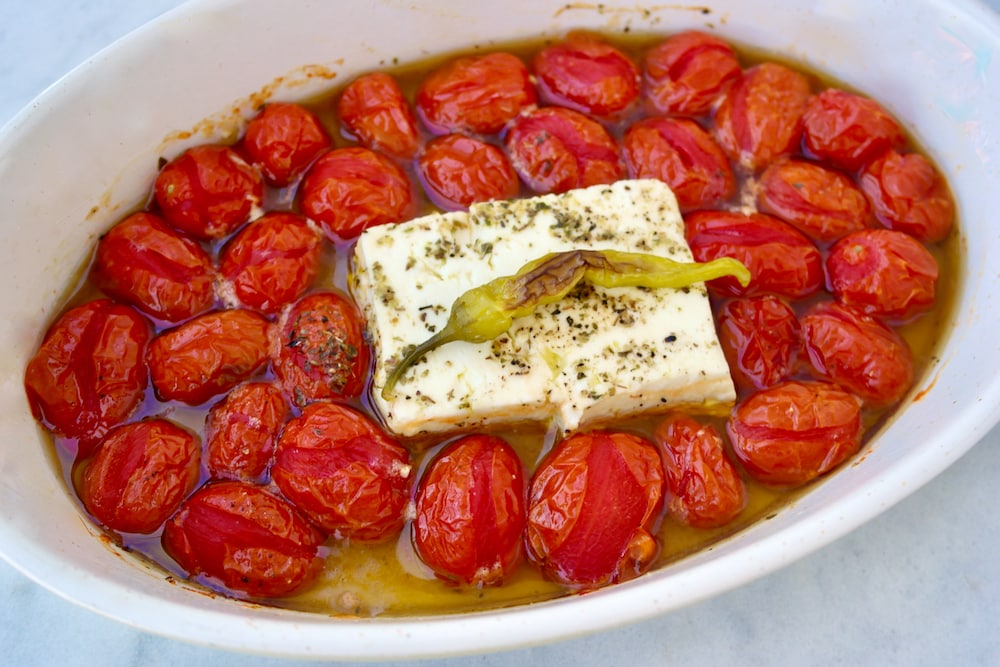 A block of feta centered in a baking dish surrounded by cherry tomatoes and garlic, with olive oil being drizzled over the top