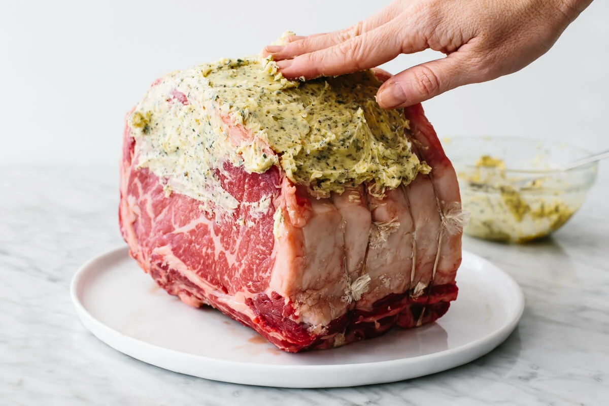 A boneless ribeye roast on a rimmed baking sheet as garlic herb butter is spread evenly over the surface