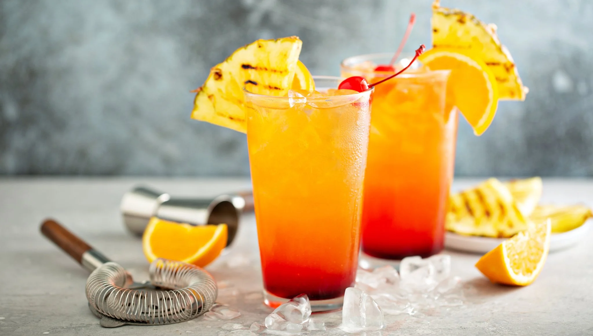 A bottle of blanco tequila, a glass of orange juice, and a small bottle of grenadine on a kitchen counter next to a cutting board with orange slices