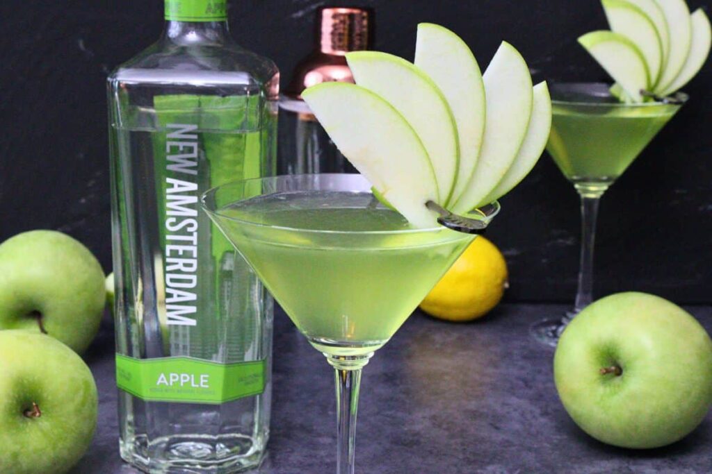 A bottle of sour apple liqueur next to vodka and a halved lemon on a kitchen counter with a small jigger