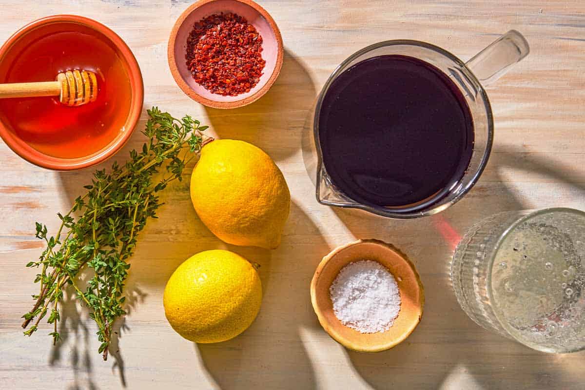 A bottle of tart cherry juice next to a halved lemon, a small bowl of salt, and a glass with ice on a neutral kitchen counter