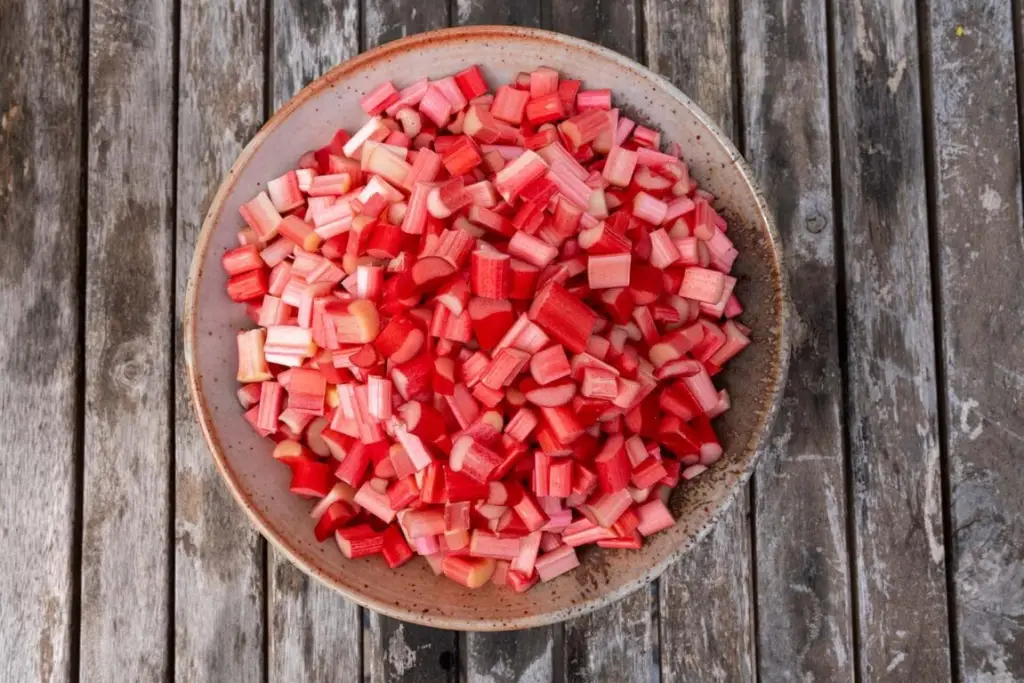 A bowl filled with evenly chopped rhubarb pieces on a kitchen counter next to a knife and cutting board