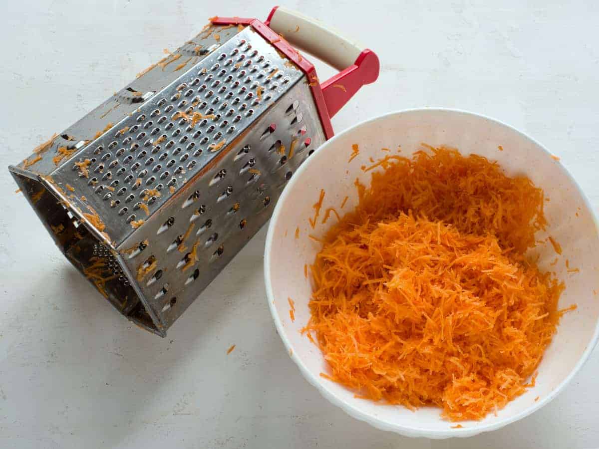 A bowl filled with finely grated carrots on a kitchen counter beside a box grater