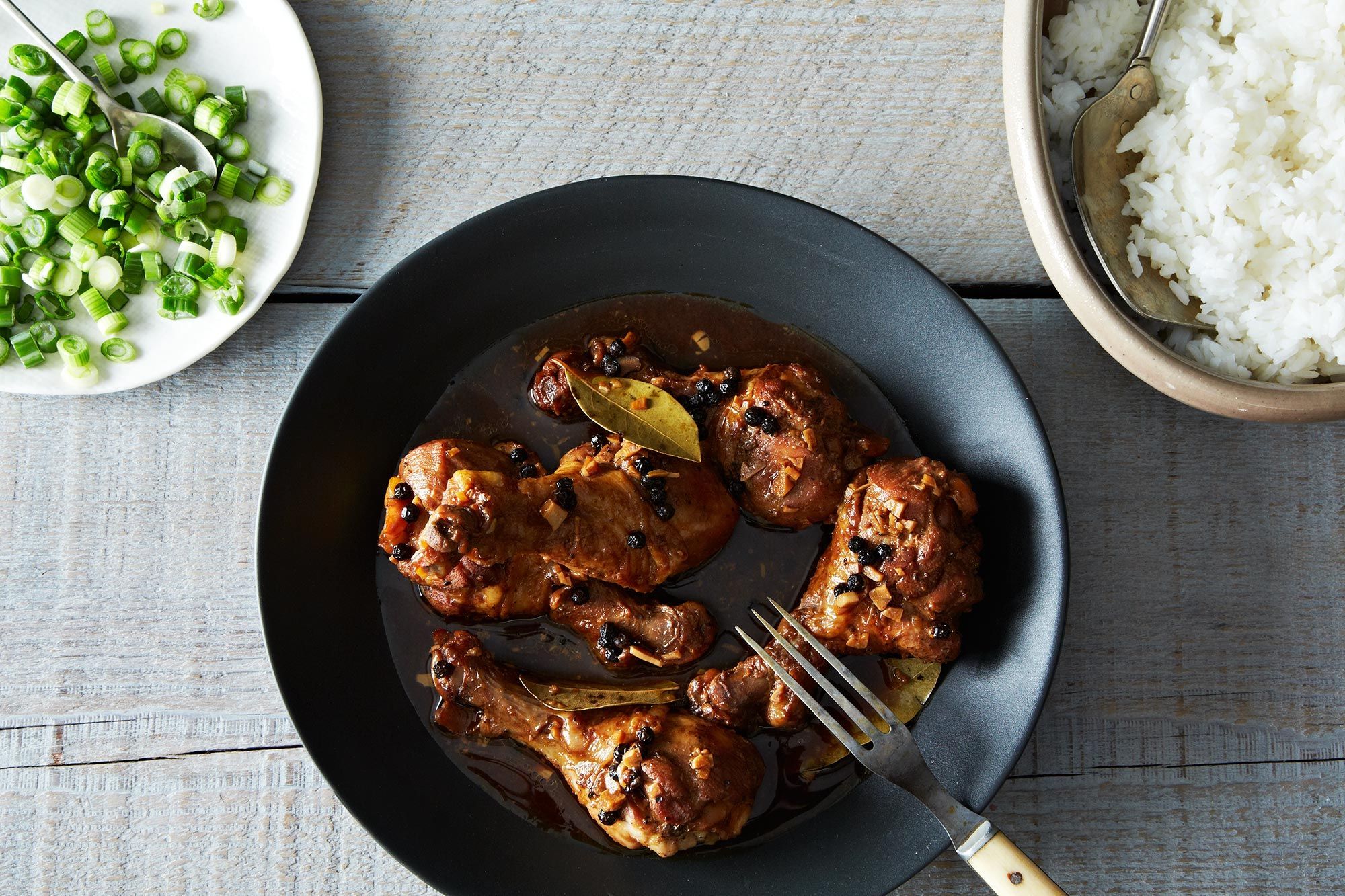 A bowl of Filipino chicken adobo with glossy dark sauce, bay leaves, and cracked pepper over steamed white rice on a warm-toned kitchen table