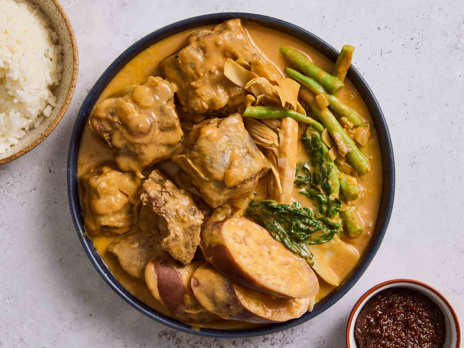 A bowl of Filipino kare kare with tender beef, eggplant, long beans, and bok choy in a creamy peanut sauce, served with rice on a warm wooden table