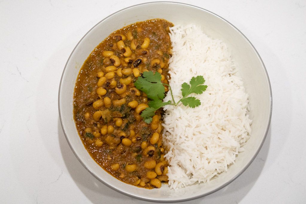 A bowl of Punjabi-style chole with chickpeas in a deep reddish-brown gravy, garnished with cilantro and sliced ginger, served beside basmati rice on a warm-toned kitchen table