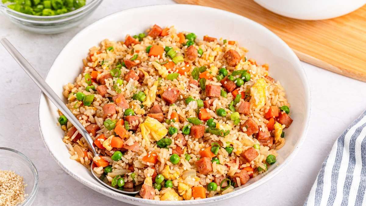 A bowl of Spam fried rice with crispy browned Spam cubes, scrambled egg, peas, and sliced scallions on a wooden table with a fork beside it