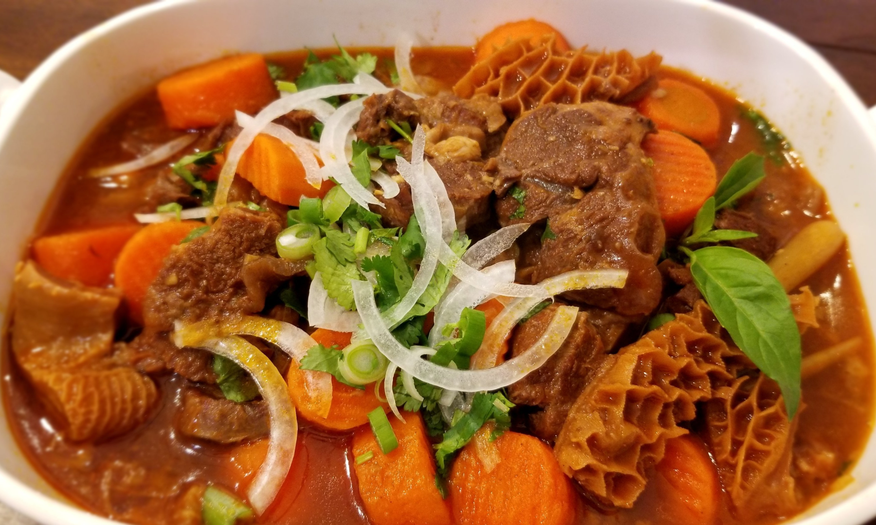 A bowl of Vietnamese bo kho beef stew with tender beef chunks, orange carrots, and a glossy reddish broth, served with fresh herbs on a rustic kitchen table