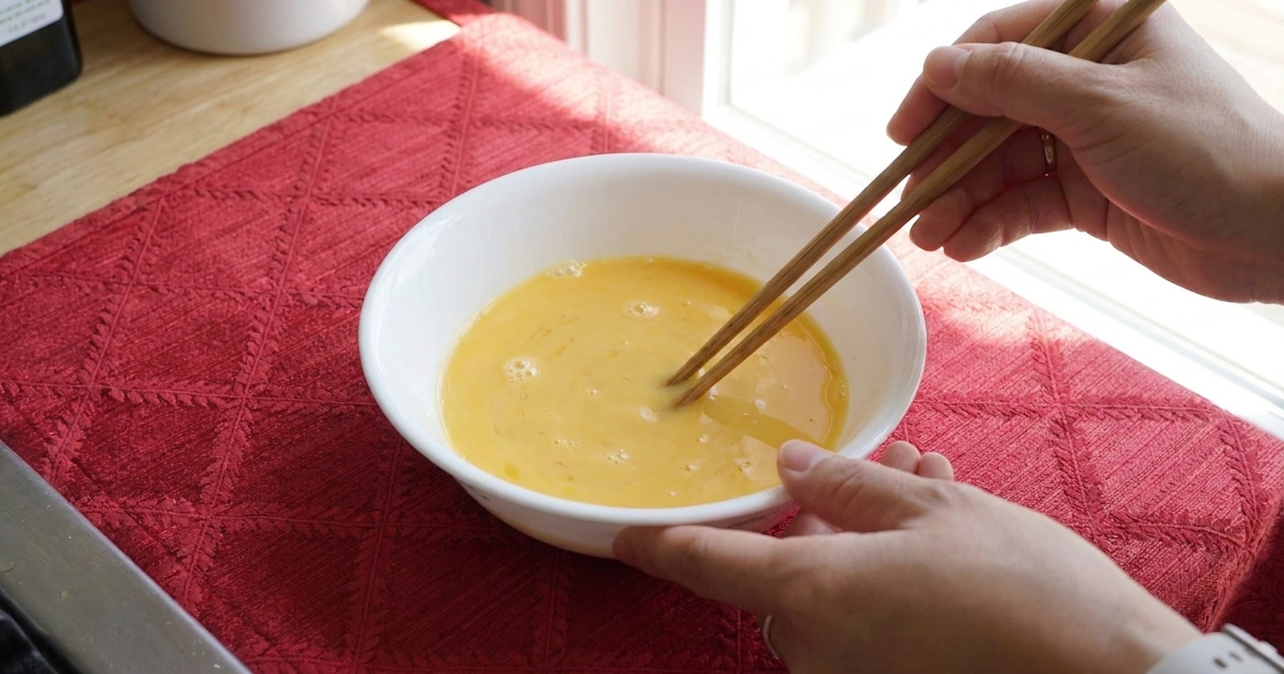A bowl of beaten eggs being stirred gently with chopsticks, with minimal bubbles on the surface