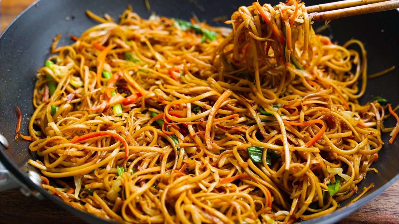 A bowl of chaomin stir-fried noodles with shredded cabbage, carrots, scallions, and glossy soy-garlic sauce on a wooden table with chopsticks