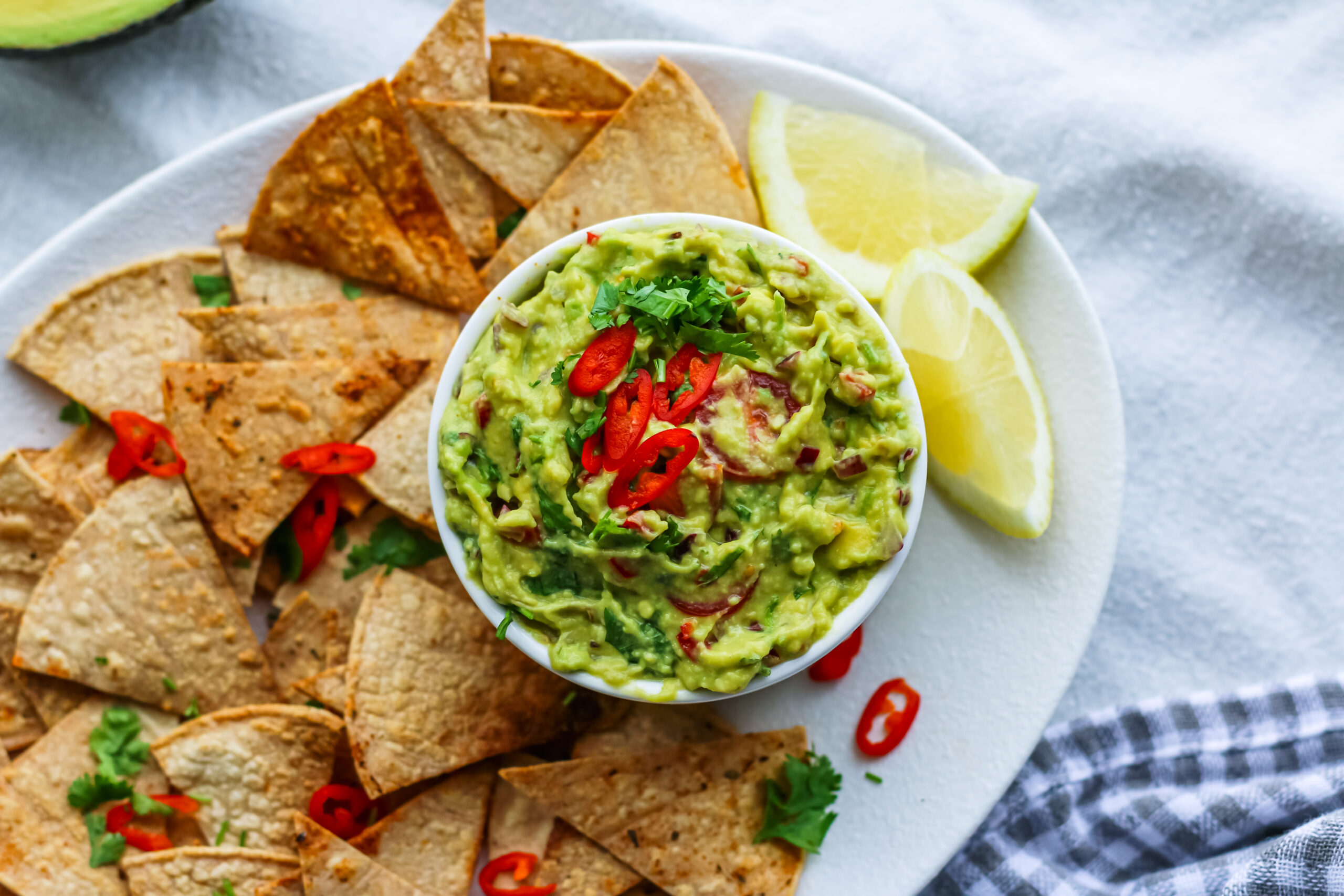 A bowl of chipotle guacamole being scooped with a tortilla chip at a casual table setting with a small plate and napkin